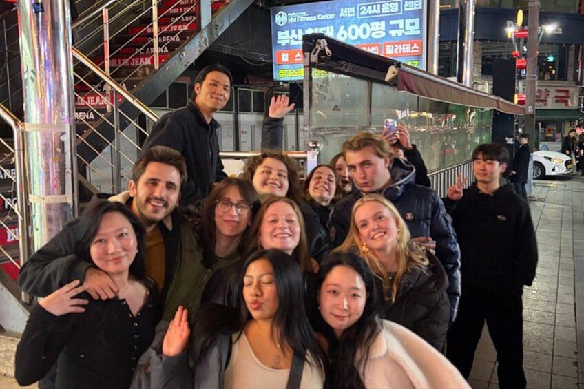 A group of people posing for a photo in an urban setting with a staircase and a building with signage in the background. 4