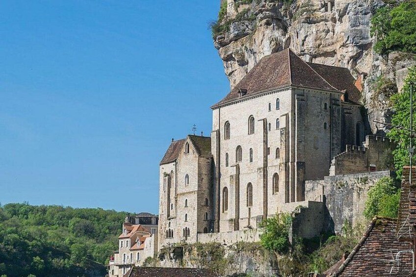 Un bâtiment historique en pierre, doté d’un toit à forte pente, situé sur une falaise, avec une végétation luxuriante en contrebas. 3