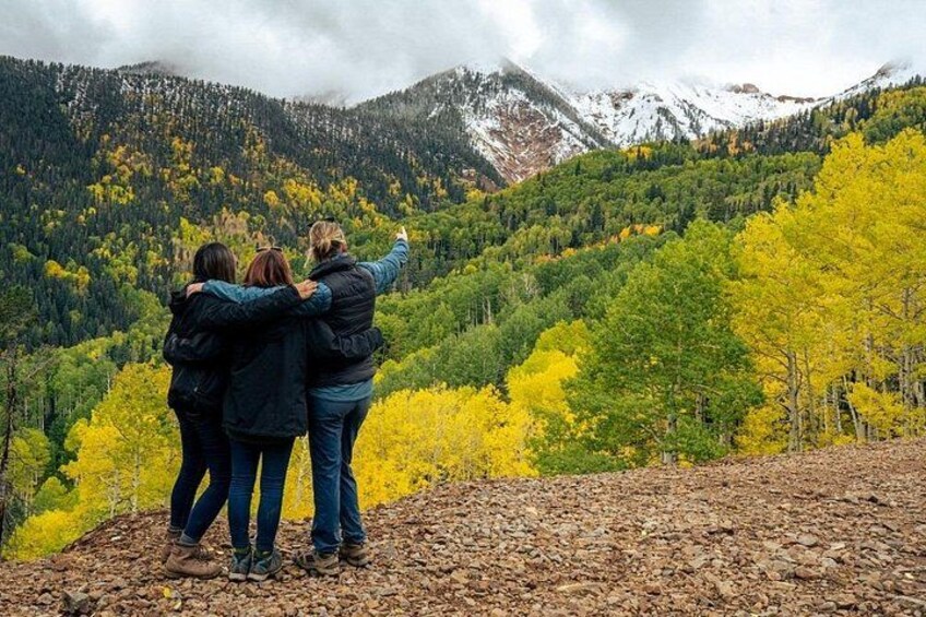 Three people standing on a rocky outcrop, overlooking a forested mountain range with autumn foliage. 3