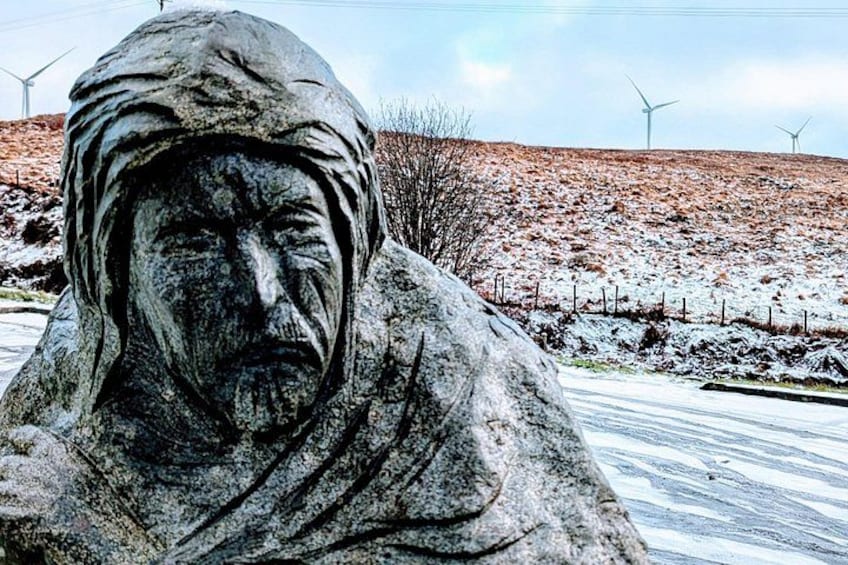 Una estatua de piedra de una persona con un tocado, sobre un fondo de paisaje montañoso con aerogeneradores. 2