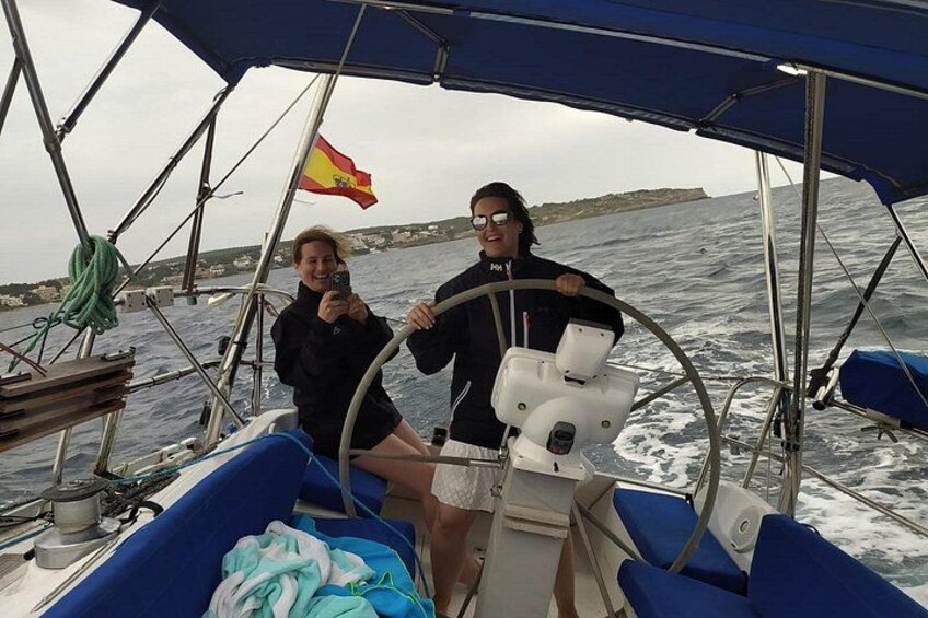 Een zeilboot met twee personen, één aan het roer en één die een foto maakt, op een wateroppervlak met een vlag en land in de verte. 3
