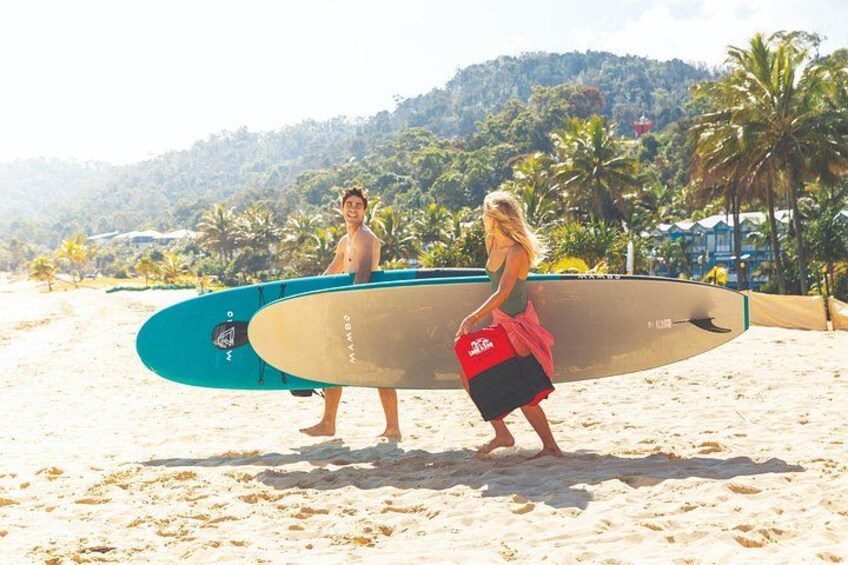 Two people on a sandy beach with a surfboard, palm trees, and a hilly background. 1