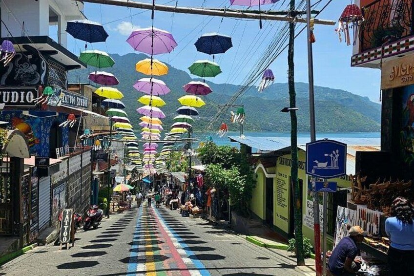 Eine Straße mit bunten Sonnenschirmen, die darüber hängen, Geschäfte an beiden Seiten und ein Blick auf das Meer im Hintergrund. 2