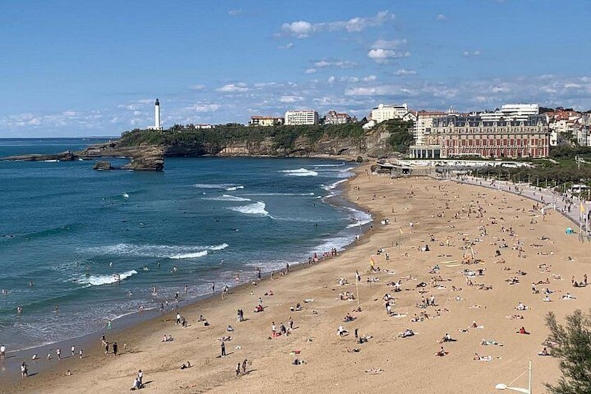 A beach with people sunbathing and swimming, a lighthouse, and buildings along the coastline. 5