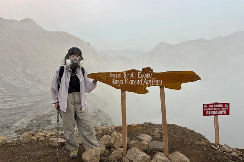 A person standing next to a wooden signpost with text, in a mountainous area with rocky terrain. 1