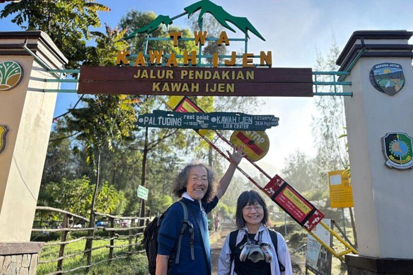 A sign with 'Kawah Ijen' and 'Jalur Pendakian Kawah Ijen' text, a wooden fence, and a person with a backpack. 3