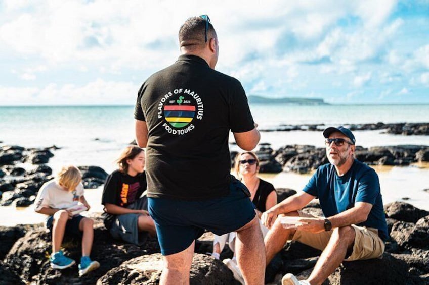 En gruppe mennesker sidder på sten ved havet, med en mand i forgrunden iført t-shirt med et logo. 1