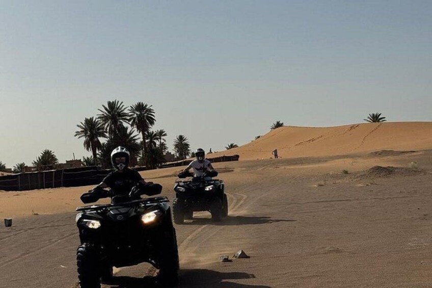 Twee personen rijden op quadbikes over een woegendheidsweg, met palmbomen en zandduinen op de achtergrond. 2