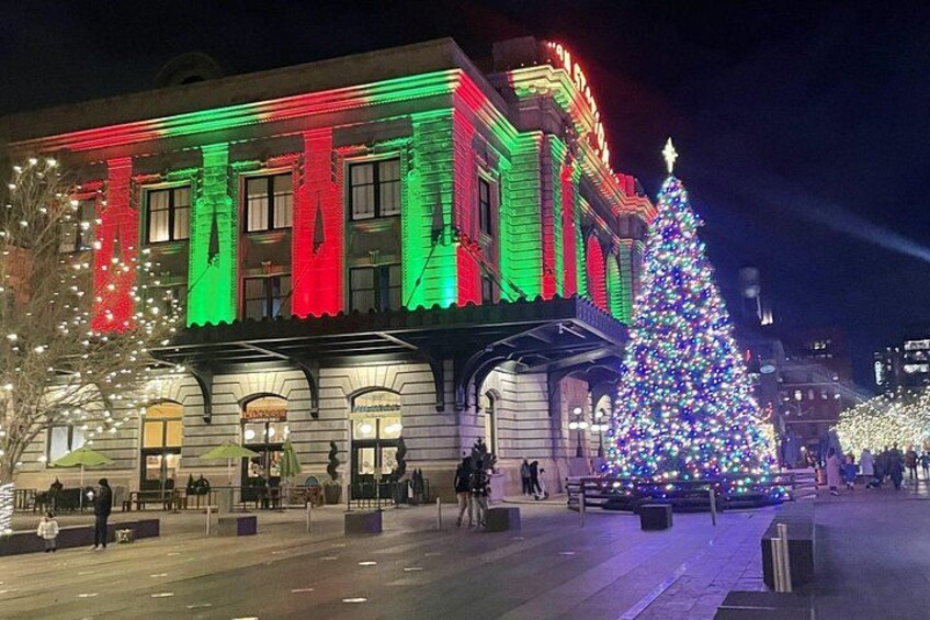 Ein Gebäude mit beleuchteten roten und grünen Lichtern, eine große Weihnachtstanne und Passanten auf dem Bürgersteig. 2