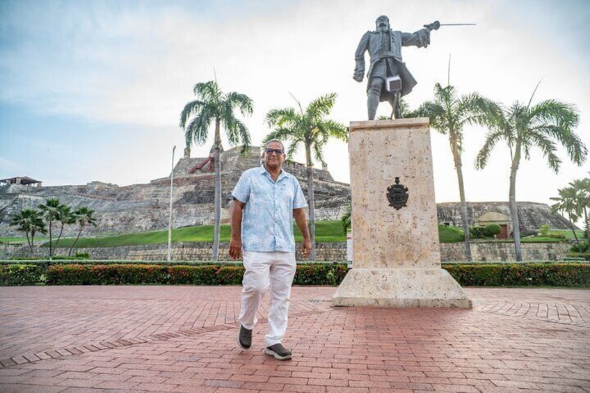 Un hombre de pie frente a una estatua sobre un sendero de ladrillos, con palmeras y una colina rocosa al fondo. 1