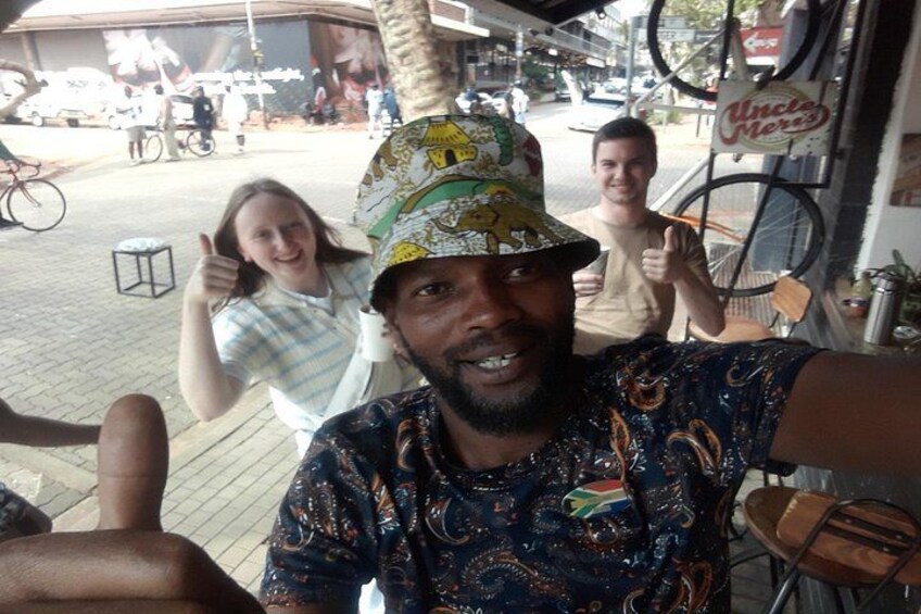 En person med en skjorta med mönster och en hatt, tillsammans med en cykel, och i bakgrunden syns en skylt med texten ”Onkel Mox”. 1