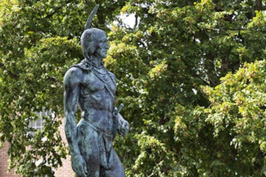 A bronze statue of a warrior holding a spear, set against a backdrop of lush green trees. 3