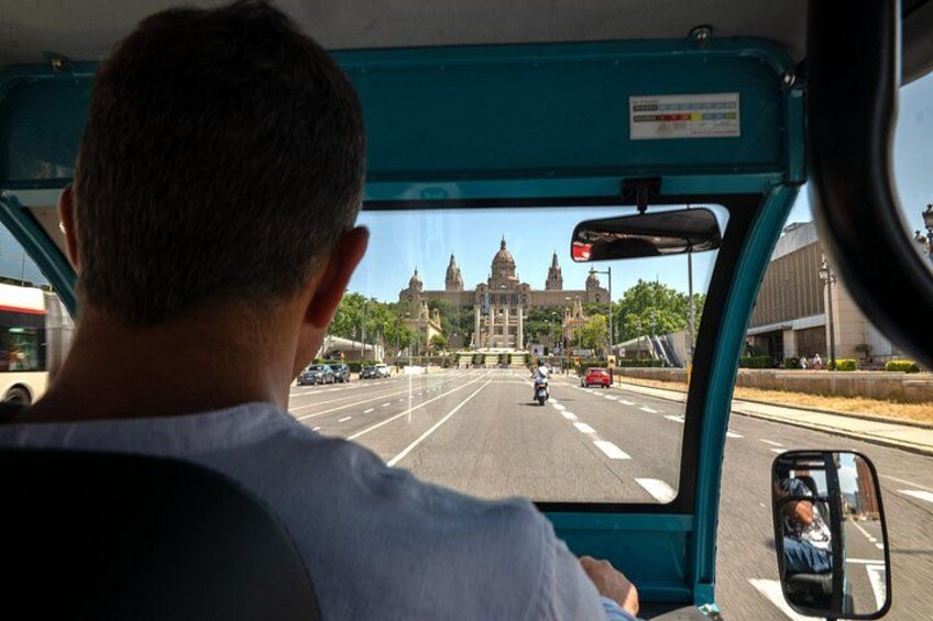 A person driving a bus, with a view of a large building in the distance. 3