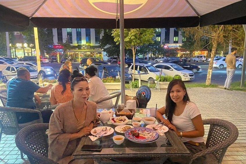 Personas cenando en un café al aire libre con mesas y sillas, una calle con coches y edificios al fondo. 5