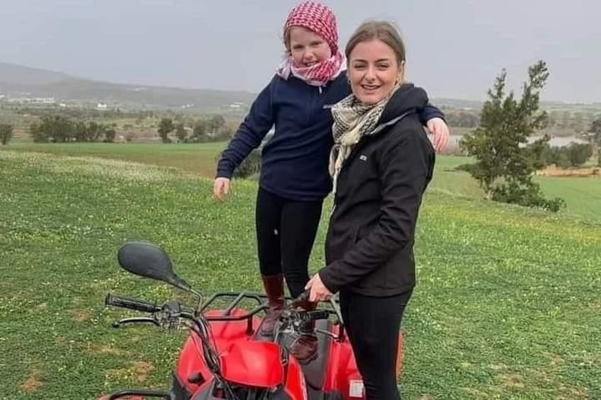 Twee personen staan naast een rode quad in een grasveld, met bomen en heuvels op de achtergrond. 2