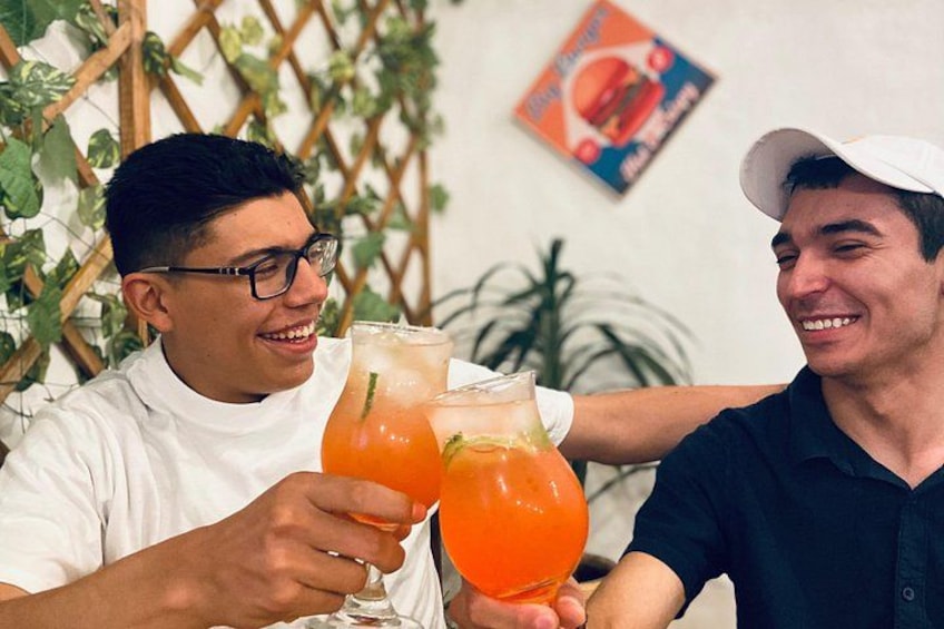 Two people holding glasses of orange beverage, a wooden lattice wall, and a poster in the background. 1