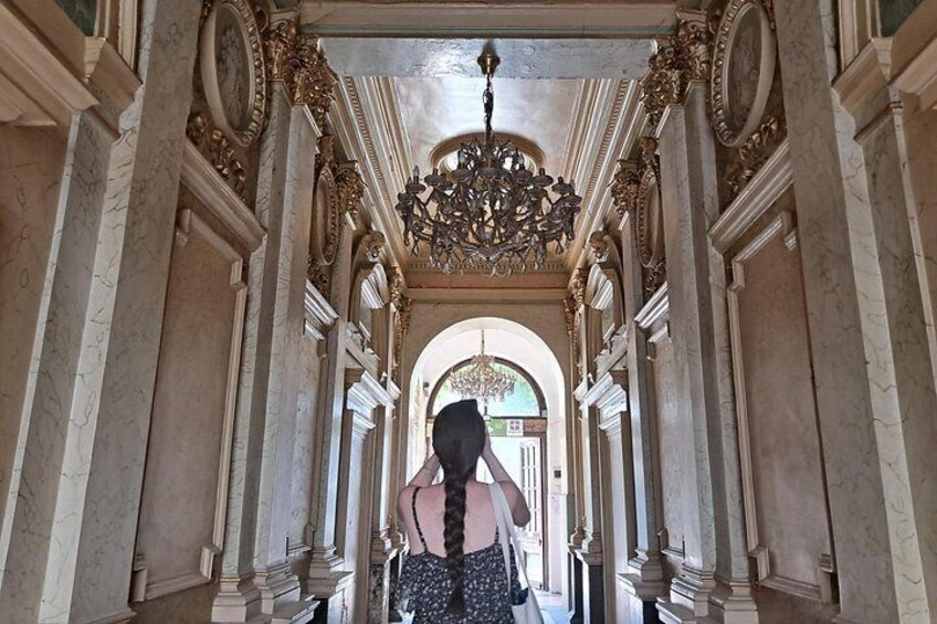 A grand hallway with ornate columns, a chandelier, and a person standing in the center. 5
