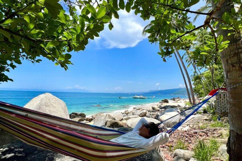 A hammock under a tree by a rocky beach with clear blue water. 2