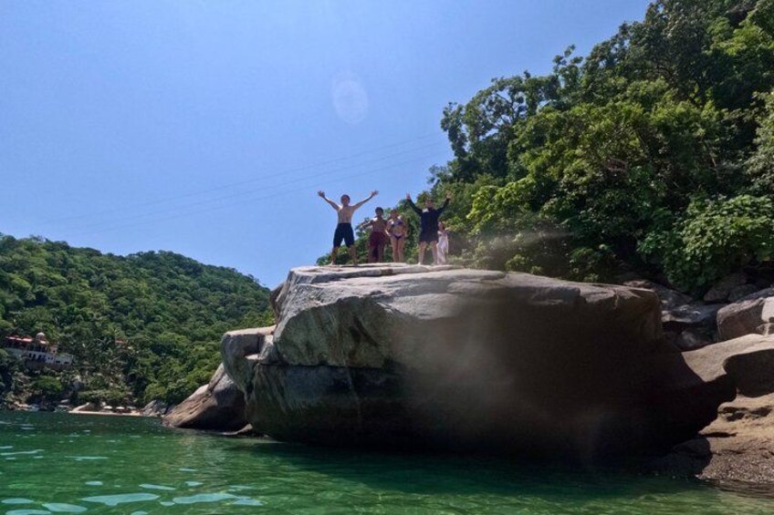 A group of people jumping off a large rock into a body of water surrounded by lush greenery. 4