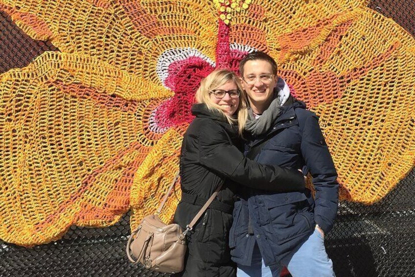 Two people hugging in front of a large, colorful crochet artwork. 2