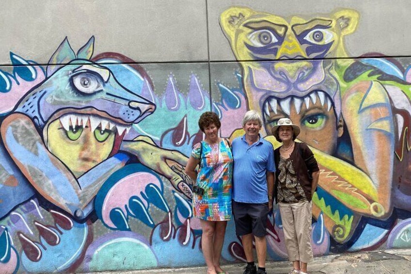 Three people standing in front of a colorful mural with abstract animal faces. 1