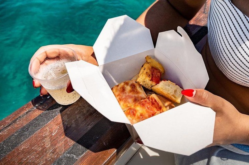 A person holding a drink and a box of food, possibly pizza, by a body of water. 2