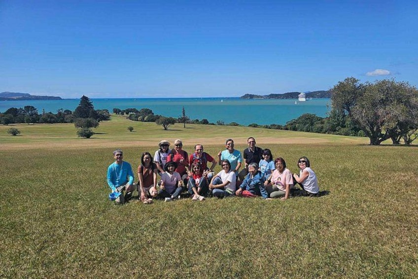 Un groupe de personnes est assis sur l’herbe dans un parc, avec une vue sur la mer et des collines en arrière-plan. 4