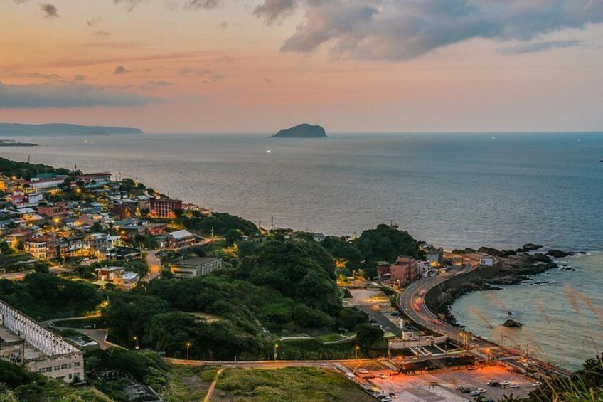 A coastal town at sunset with buildings, a curved road, and a distant island. 1