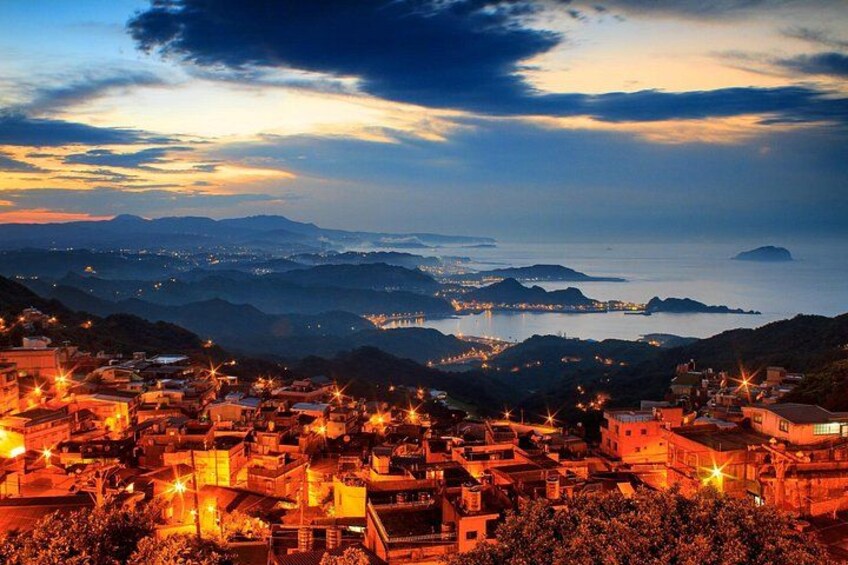 A coastal town at dusk with illuminated buildings and a view of the sea and distant mountains. 3