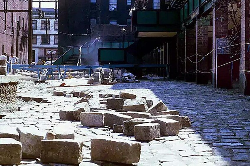 A cobblestone street with large stone blocks in the foreground, a building with a green facade in the background, and a staircase leading up to a second floor. 3