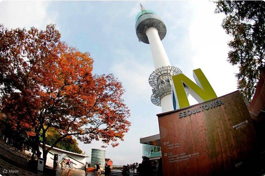 A tower with a spherical observation deck, a large 'N' sign, autumn-colored trees, and a modern building with glass windows. 1