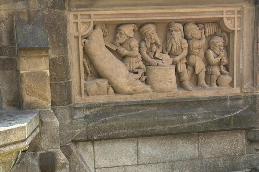 Een stenen beeldhouwwerk van vier figuren, één zittend en drie staand, met een groot dier en een pot. 3