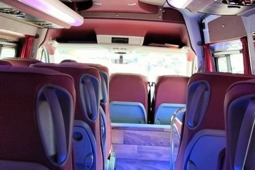 El interior de un autobús con asientos rojos y azules, un pasillo central y compartimentos superiores para equipaje. 2