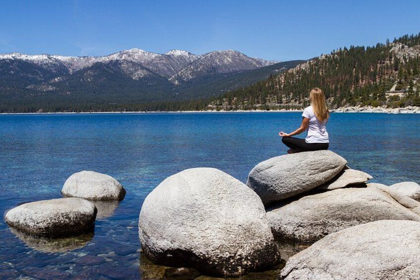 A person meditating on rocks by a clear lake with mountains in the background. 5
