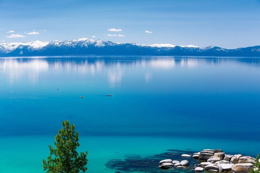 A calm lake with clear blue water, a tree in the foreground, and snow-capped mountains in the background. 3