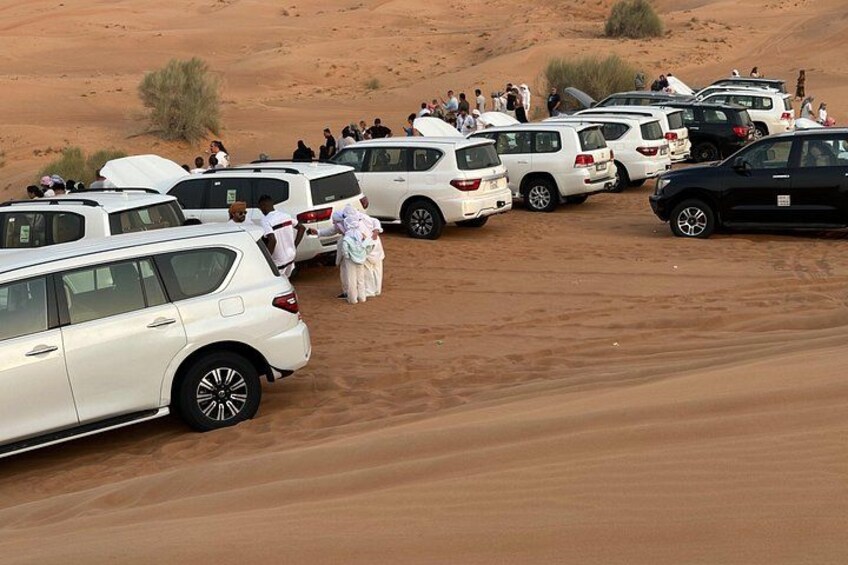 A desert with white cars parked in a line, people standing around, and a few individuals in traditional attire. 1