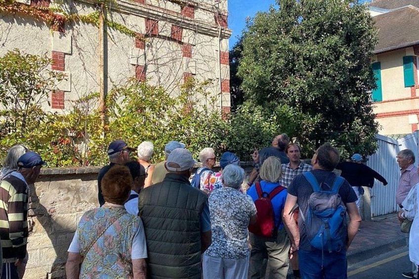 Un grupo de personas de pie al aire libre, algunas con mochilas, cerca de un edificio de fachada de ladrillo y un árbol al fondo. 2