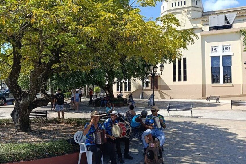 A group of people playing musical instruments under a tree in a public square. 2
