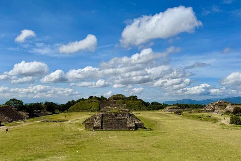 Ancient ruins with a large central pyramid and smaller structures in a grassy field under a blue sky with clouds. 3