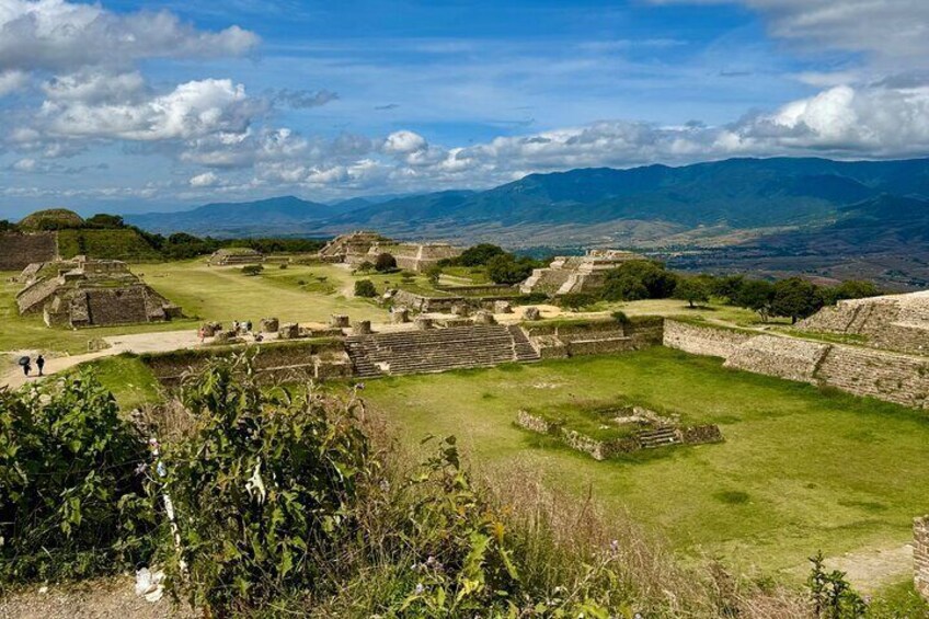 Ancient ruins with stepped pyramids and a large open plaza, surrounded by lush greenery and distant mountains. 1