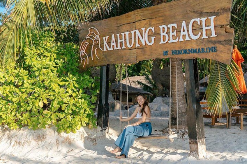 Een houten bord met daarop 'Kahung Beach' en 'by Nikornmarine', een persoon zittend op een schommel, palmbomen en een zandstrand. 5