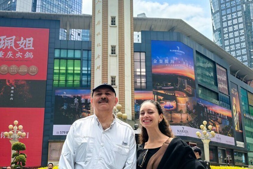 Een man en een vrouw staan voor een gebouw met grote digitale billboards en een hoge structuur op de achtergrond. 5