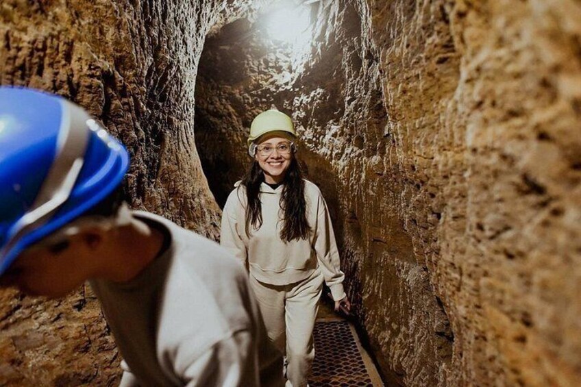 A person in a hard hat and another in a light-colored outfit in a cave with a metal walkway. 3