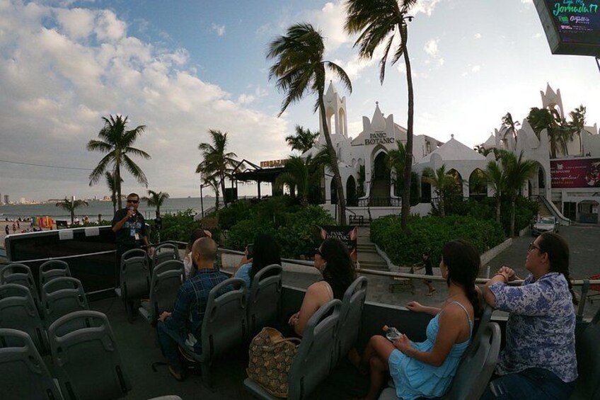 A group of people seated on a boat, a speaker addressing them, and a white building with a pointed roof in the background. 5