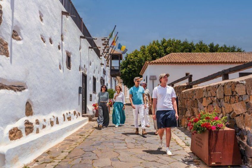 A narrow cobblestone street lined with white buildings, a stone wall, and a wooden structure with red flowers. 1