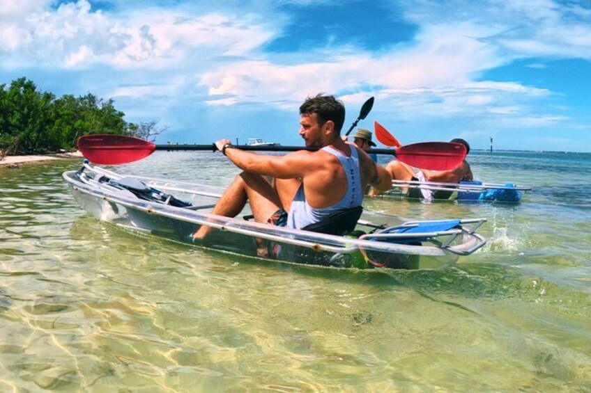 A man paddling a canoe on clear shallow water with a distant shoreline and trees. 2