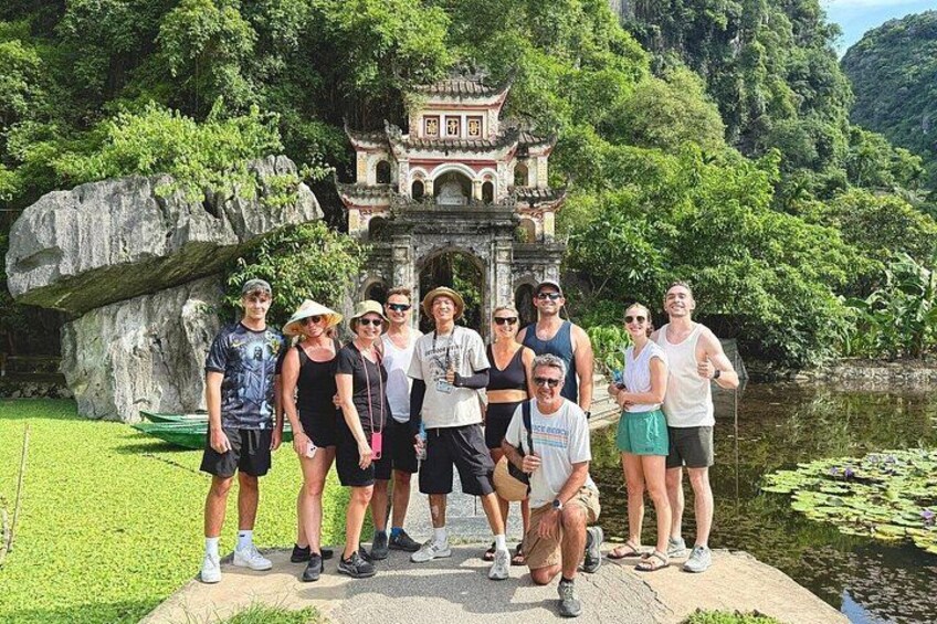 A group of people posing in front of a traditional temple with a rock formation and a pond in the background. 1