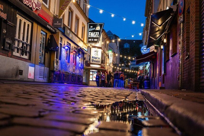 A cobblestone street at night with illuminated storefronts and a wet surface reflecting lights. 5