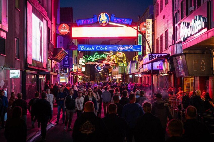 A crowded street with neon signs advertising various establishments like a dance club and a beach club. 4