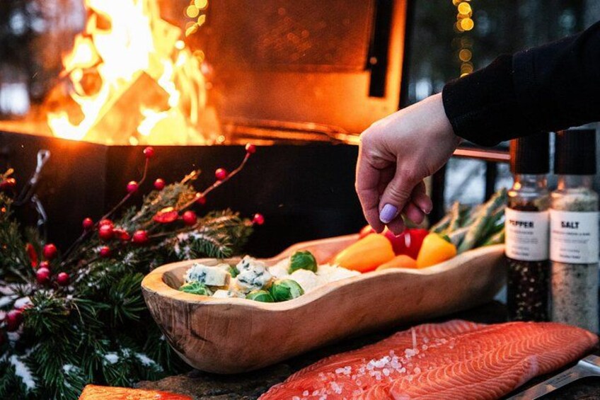 A hand seasoning salmon with salt and pepper, with a wooden bowl containing vegetables and a fire in the background. 3
